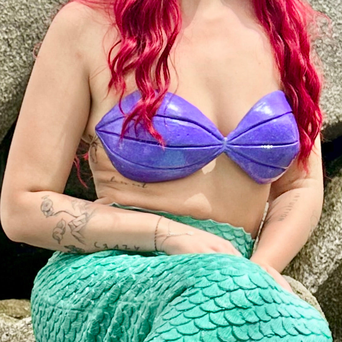 Person wearing a purple mermaid top and green tail against a stone background. Ariel Little mermaid cosplay outfit costume