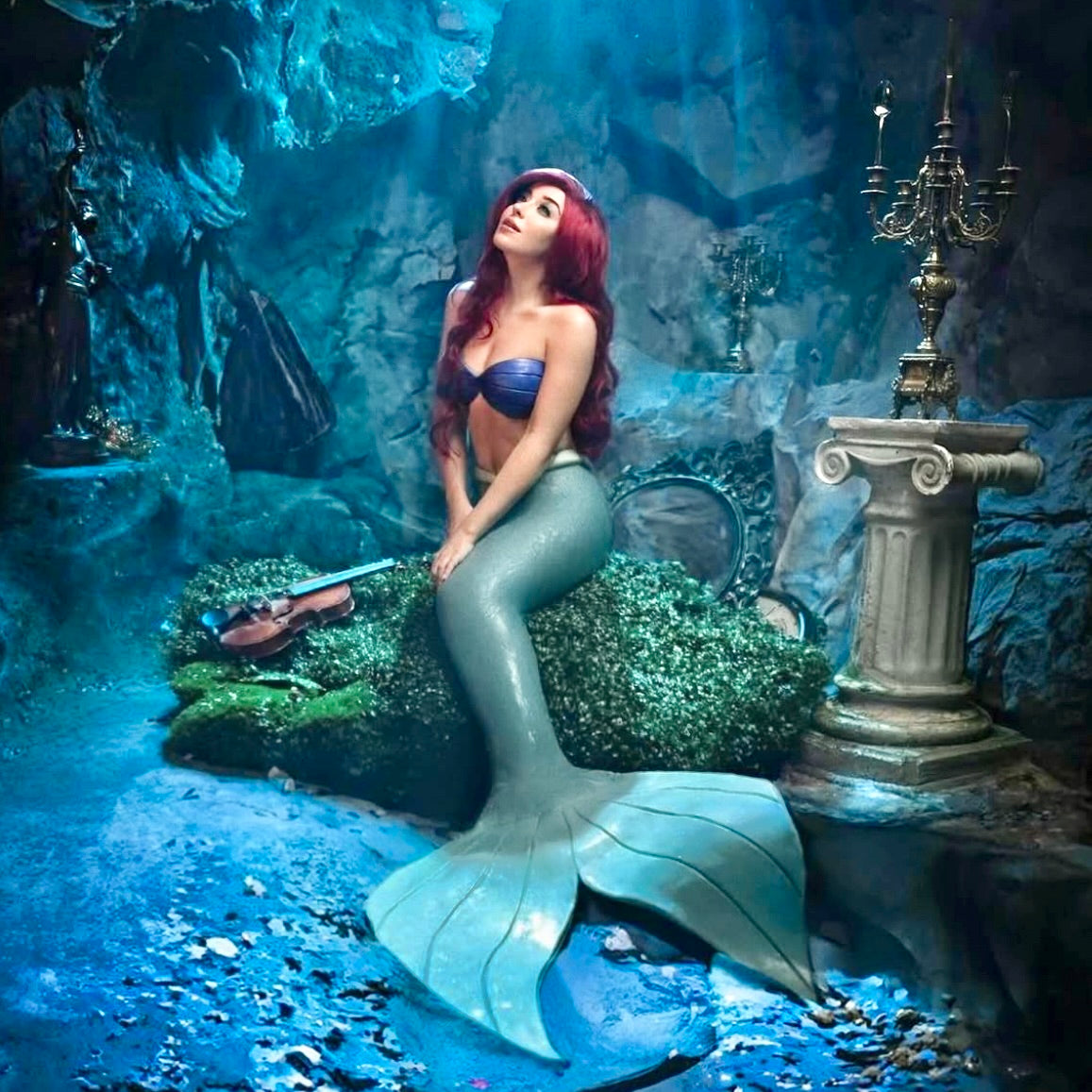 Mermaid sitting on a rock in an underwater setting with decorative elements. Custom-made Ariel mermaid top in silicone with skin-tone power mesh, swimmable, Disney-inspired cosplay bra with seashell design
