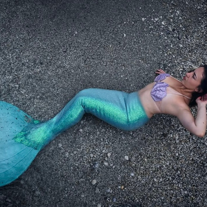 Person wearing a mermaid tail lying on a pebble beach. Green silicone mermaid tail with purple shell top