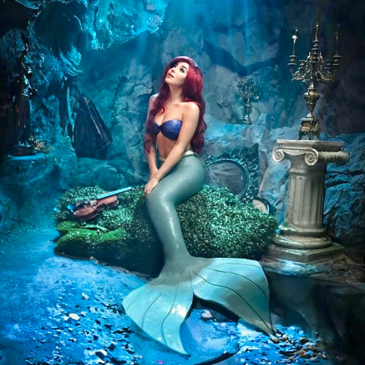 Mermaid sitting on a rock in an underwater setting with decorative elements. Custom-made Ariel mermaid top in silicone with skin-tone power mesh, swimmable, Disney-inspired cosplay bra with seashell design
