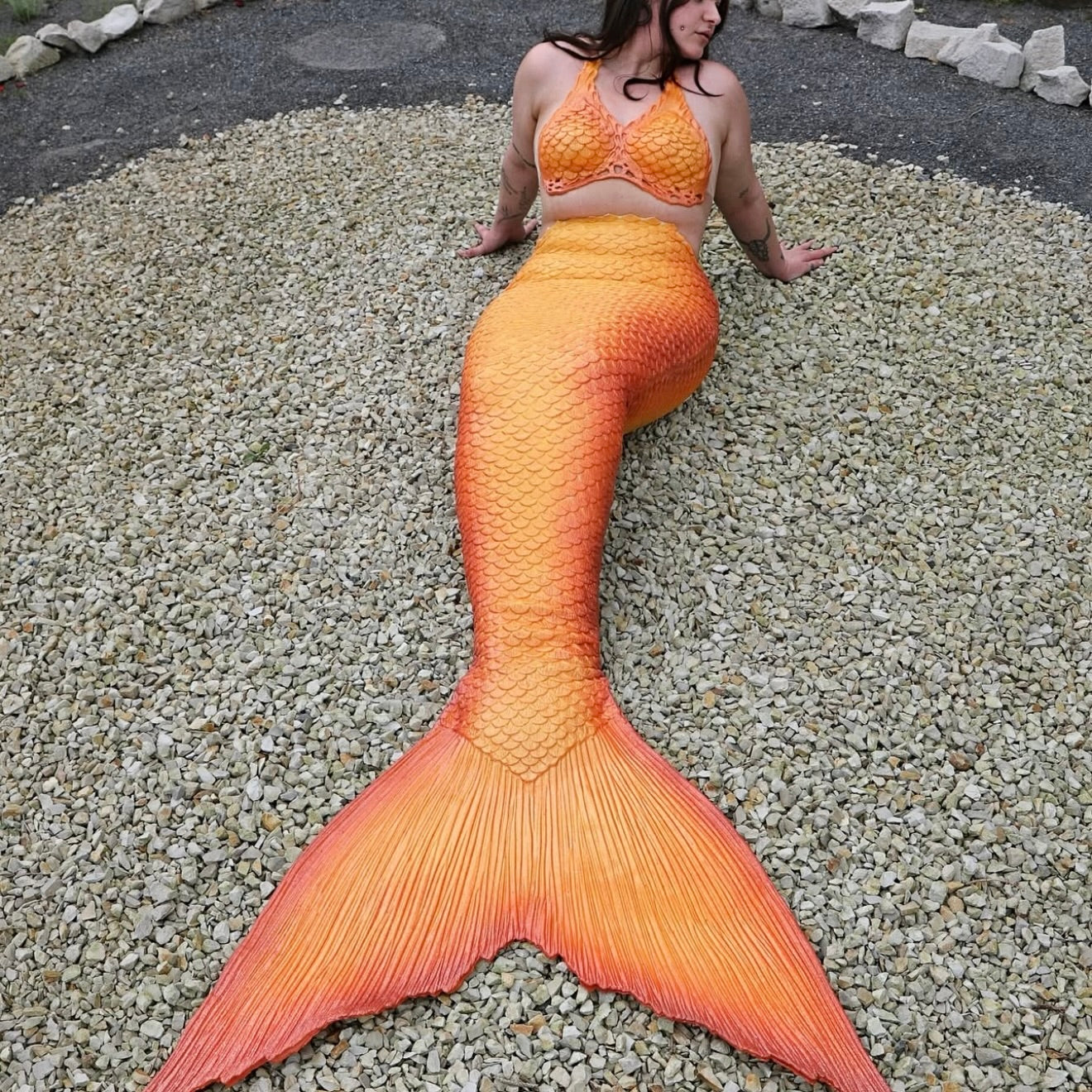 Orange silicone mermaid and mermaid top. Mako mermaid cosplay outfit costume. Person wearing an orange mermaid tail on a gravel surface