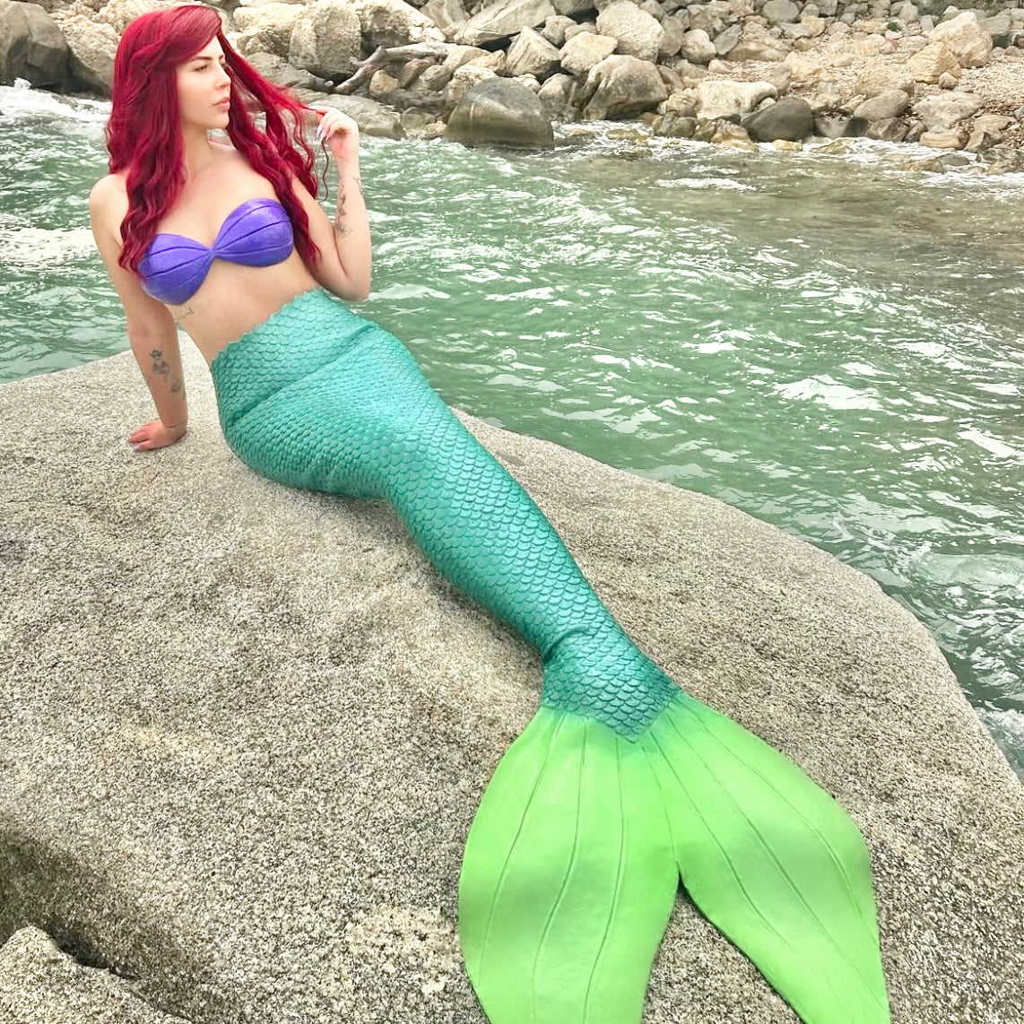 Person in a mermaid tail sitting on a rock by the water. Handmade silicone mermaid top for underwater photoshoots, Ariel Little Mermaid design